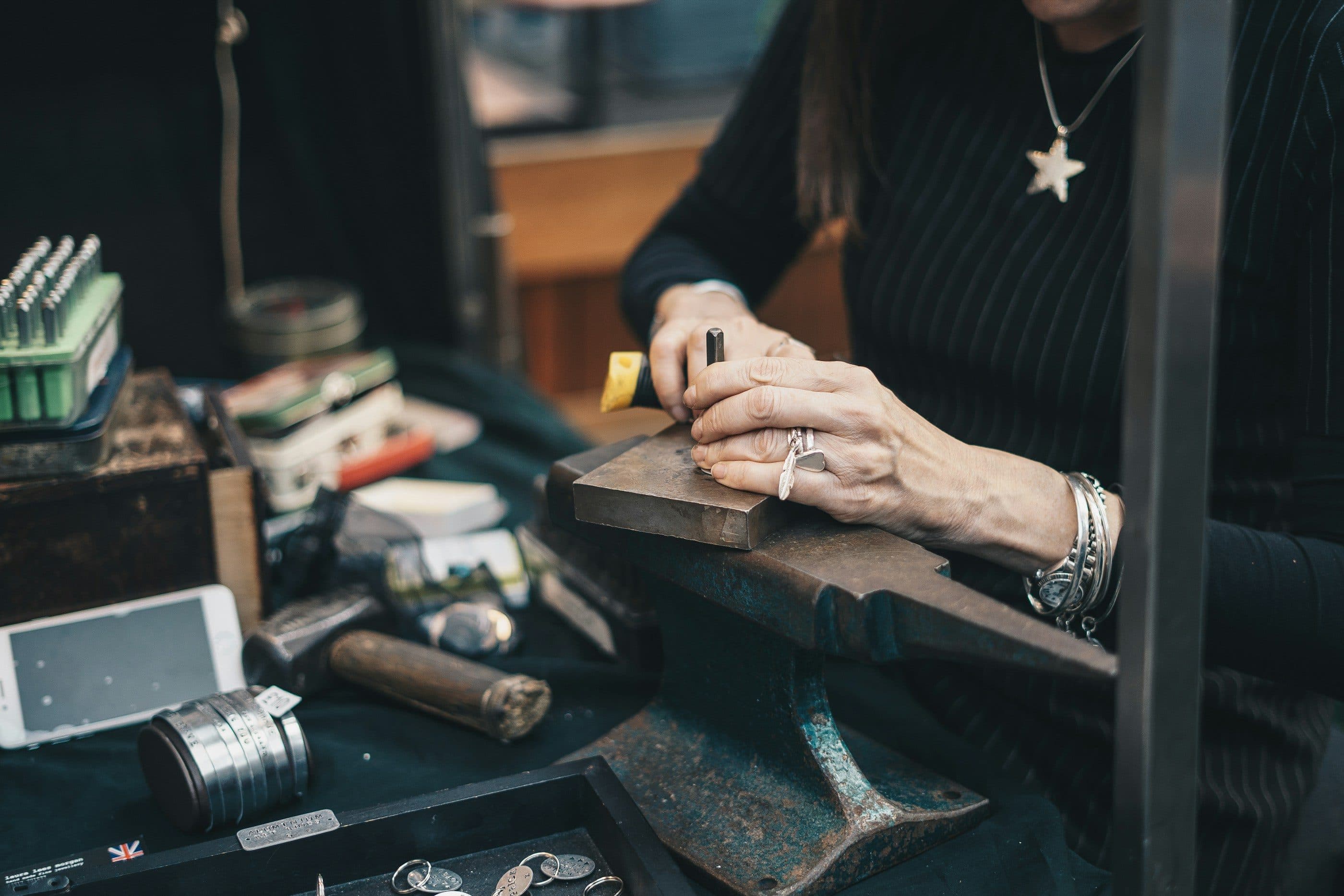 Mãos de uma artesã, com anéis e pulseiras, a trabalhar uma peça sobre a bigorna